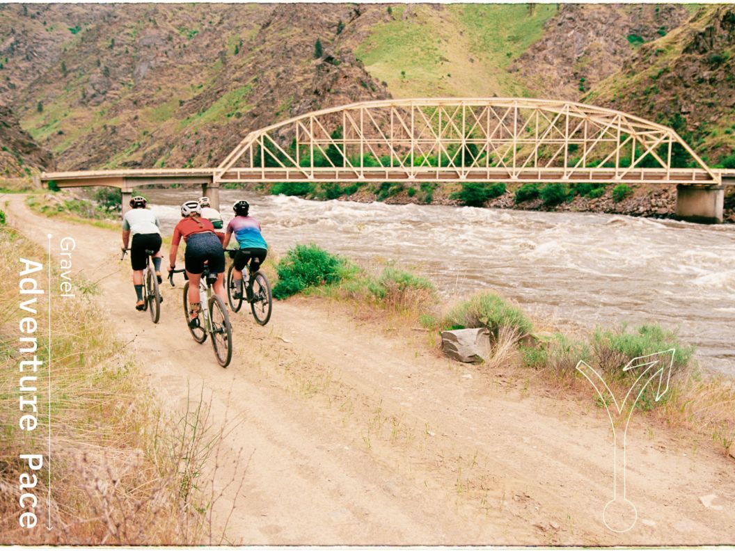 Four gravel riders pedal toward a bridge crossing fast-flowing river.